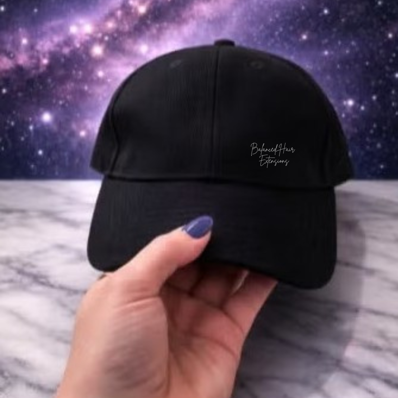 A black baseball cap with red light therapy pads inside held by a hand with purple nail polish, featuring white embroidered text on the side, set against a starry galaxy background and a gray marble surface.